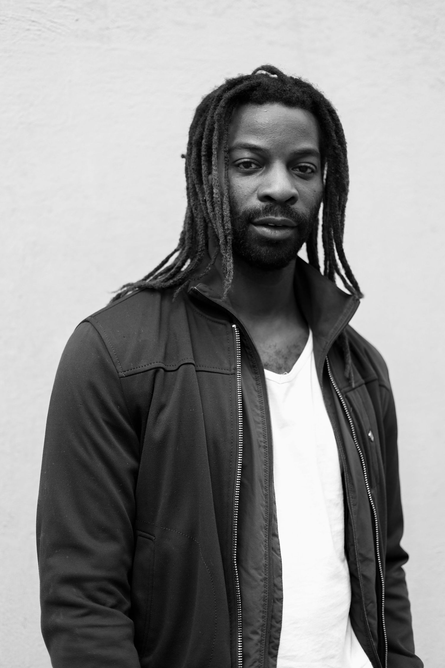 Black and white portrait featuring long dreadlocks, white shirt and black jacket against a plain wall.