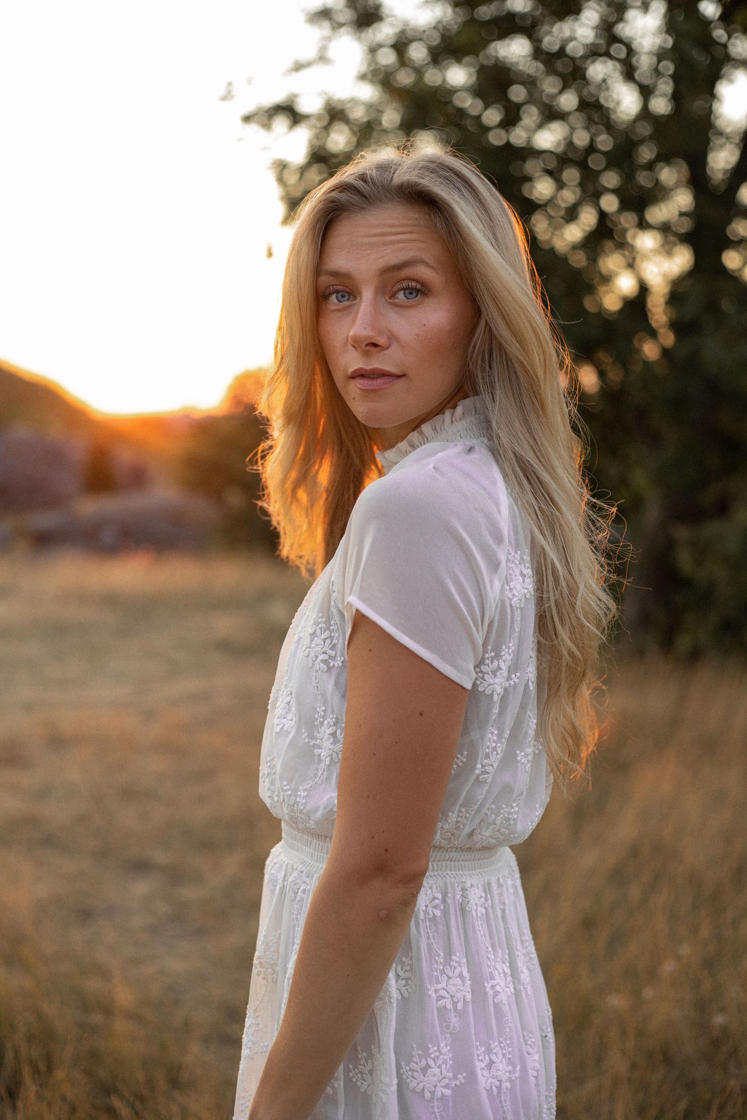 A woman in a white lace dress poses against golden sunlight in a field at sunset.