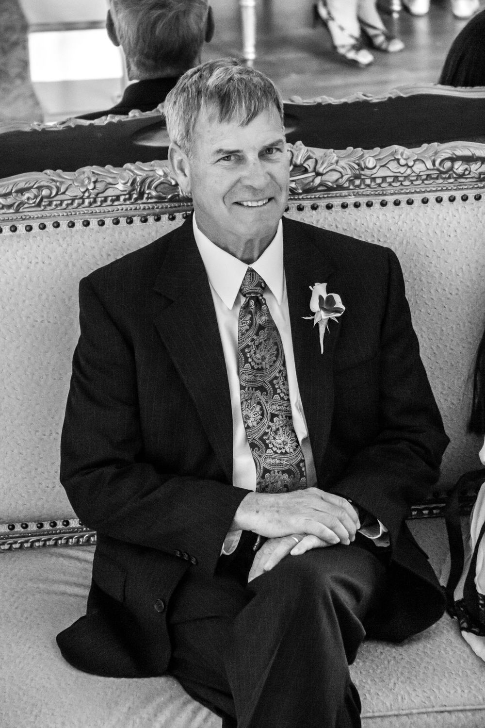 A black and white photo shows a person in a dark suit sitting on an ornate couch smiling at the camera.