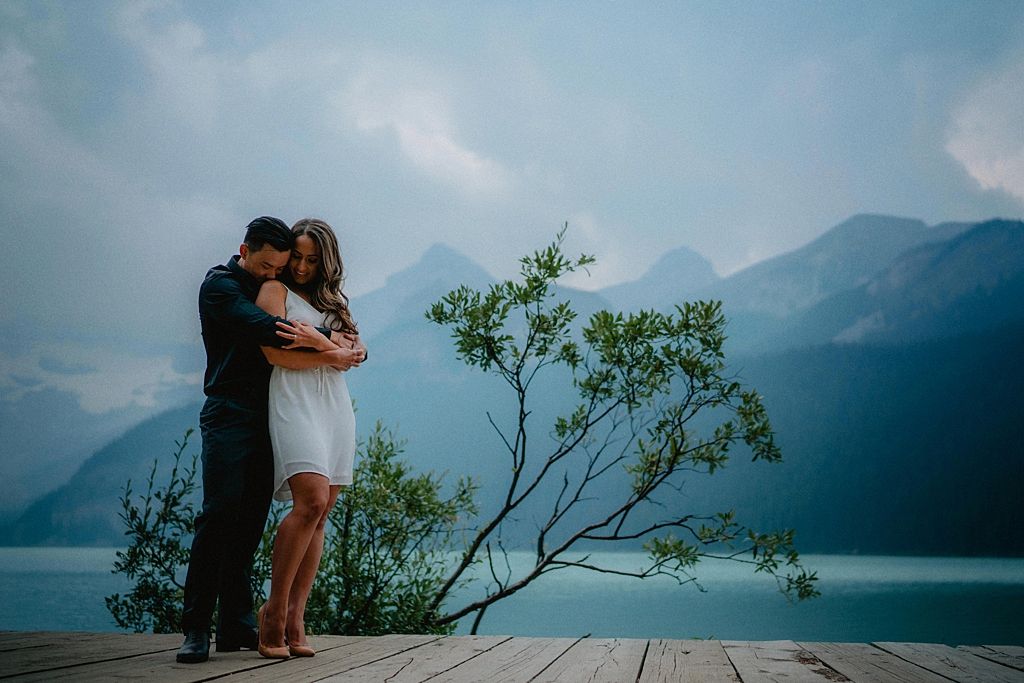 Couple embracing on wooden deck overlooking misty mountain lake with small tree.