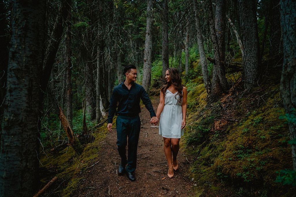 A couple walks hand in hand along a mossy forest path surrounded by tall trees.