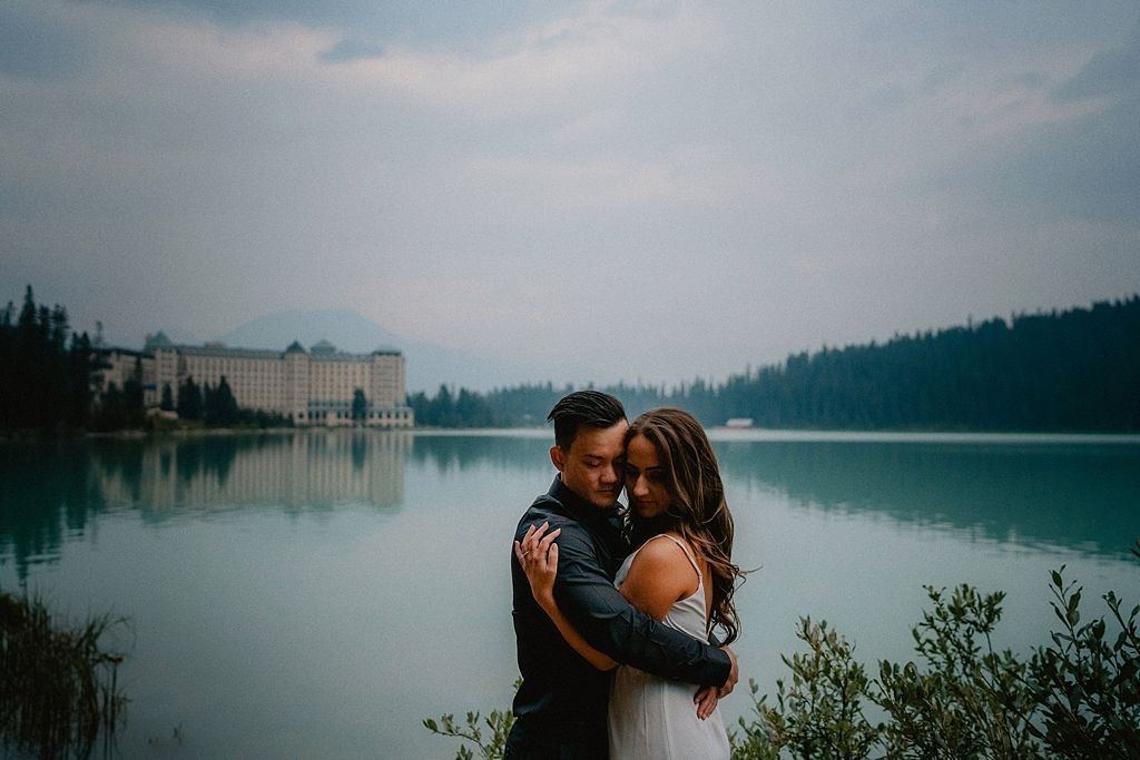 Couple embraces by a serene mountain lake with evergreen forests in the background.