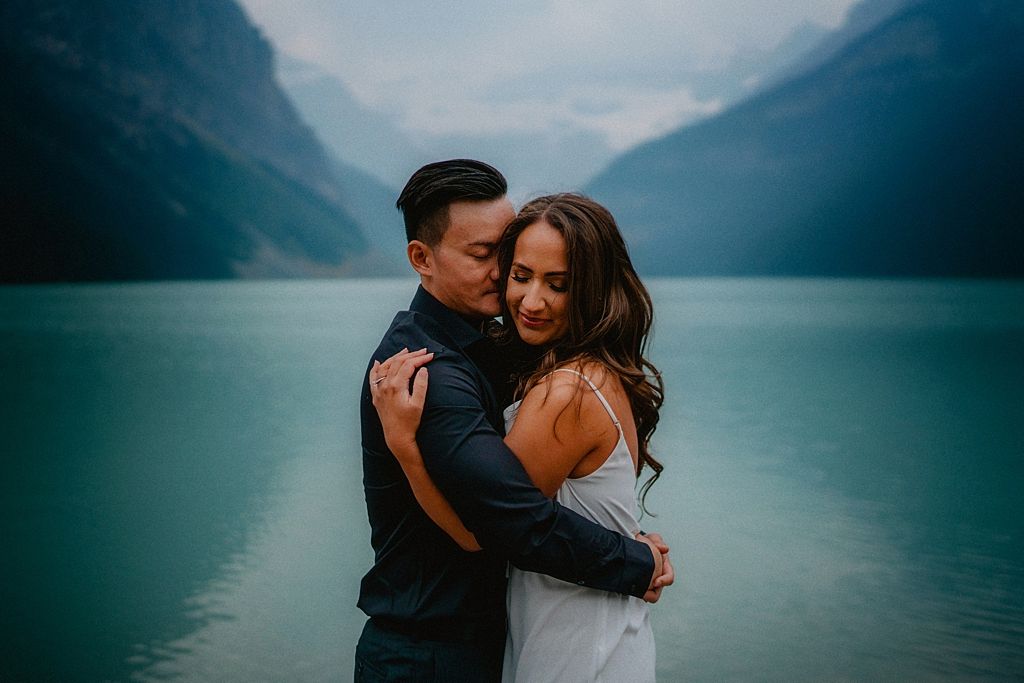 A romantic couple embraces against a backdrop of turquoise lake waters and mountain peaks in a moody setting.