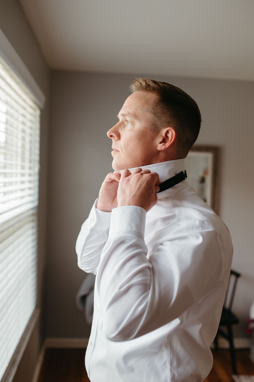 A person adjusts their bowtie while looking out a window, dressed in formal white attire.