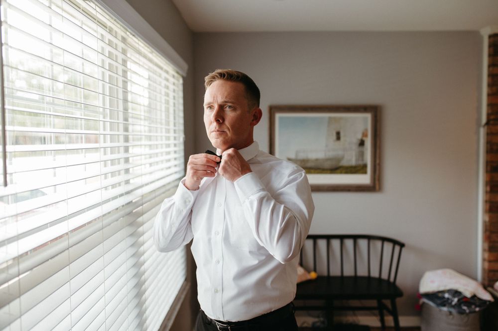 A person adjusting a bowtie while standing near venetian blinds in a formal white dress shirt.