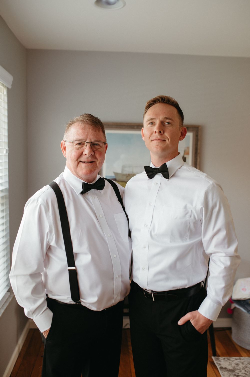 Two people in formal attire with bowties and suspenders pose in a well-lit room.