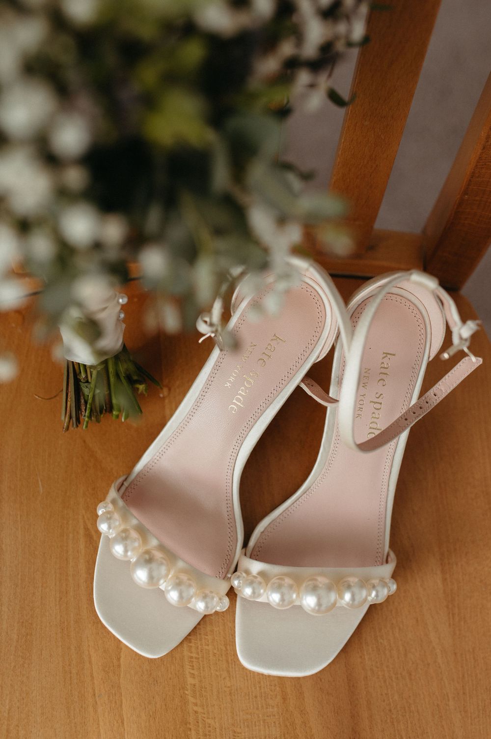 Elegant white wedding sandals with pearl details displayed on wooden surface next to flowers.