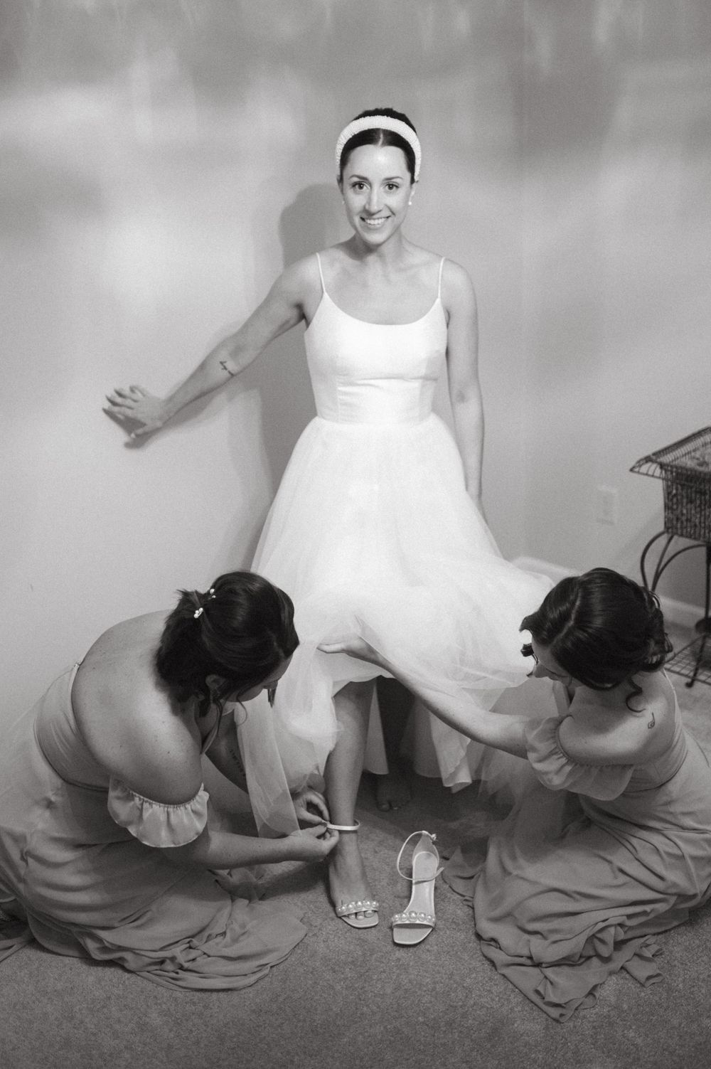Black and white photo of wedding dress preparations with helpers adjusting details.