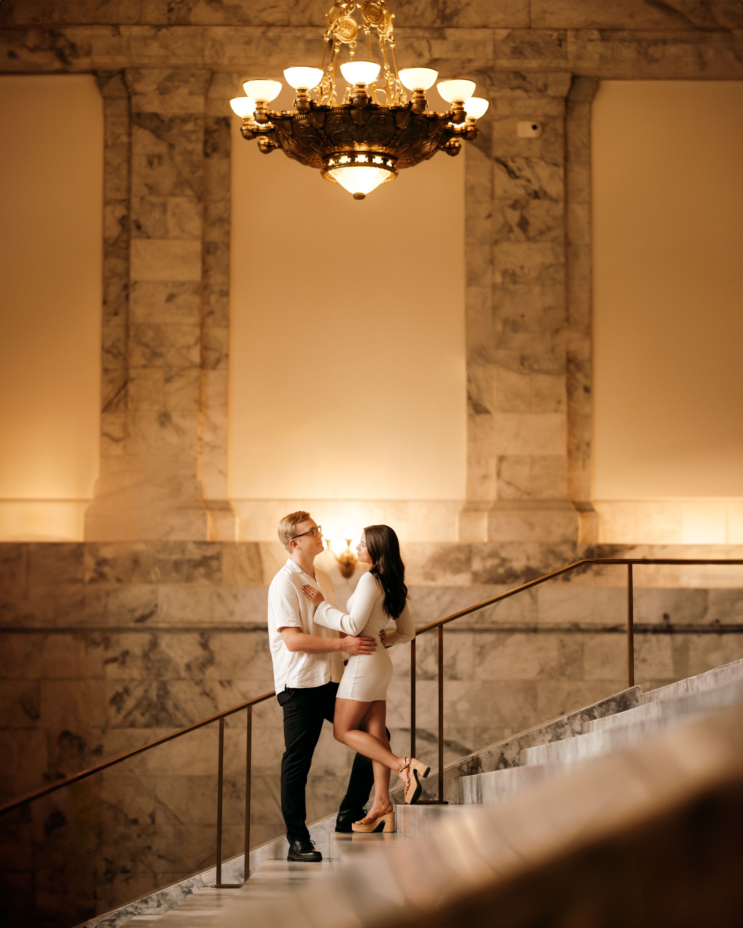 Couple shares romantic moment on marble staircase under ornate chandelier.