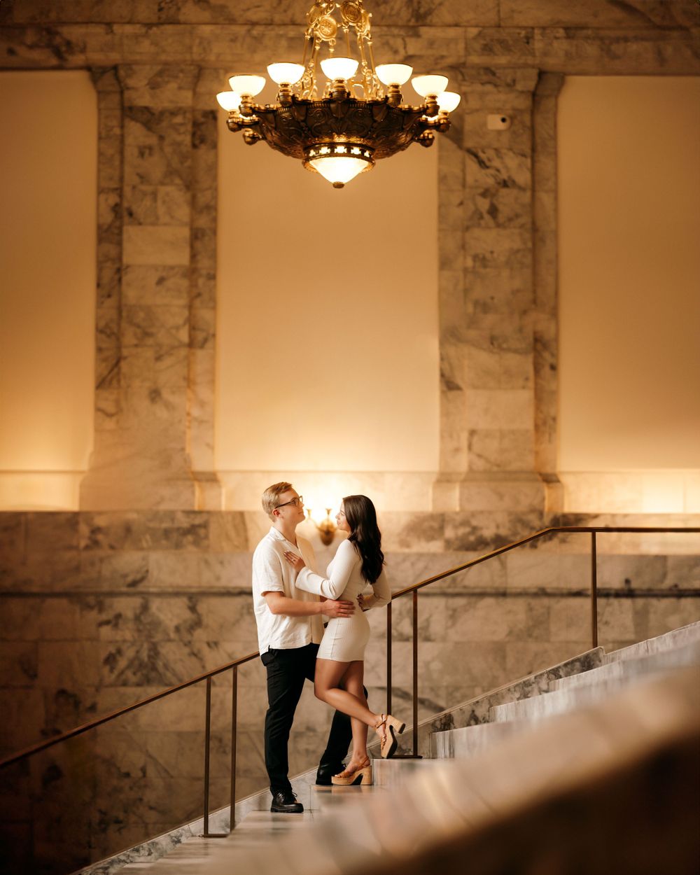 Couple shares romantic moment on marble staircase under ornate chandelier.