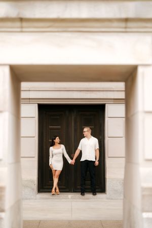 A couple stands framed in a doorway of a stone building holding hands.