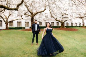 A couple in navy formal attire stands among blooming cherry blossom trees on a green lawn.