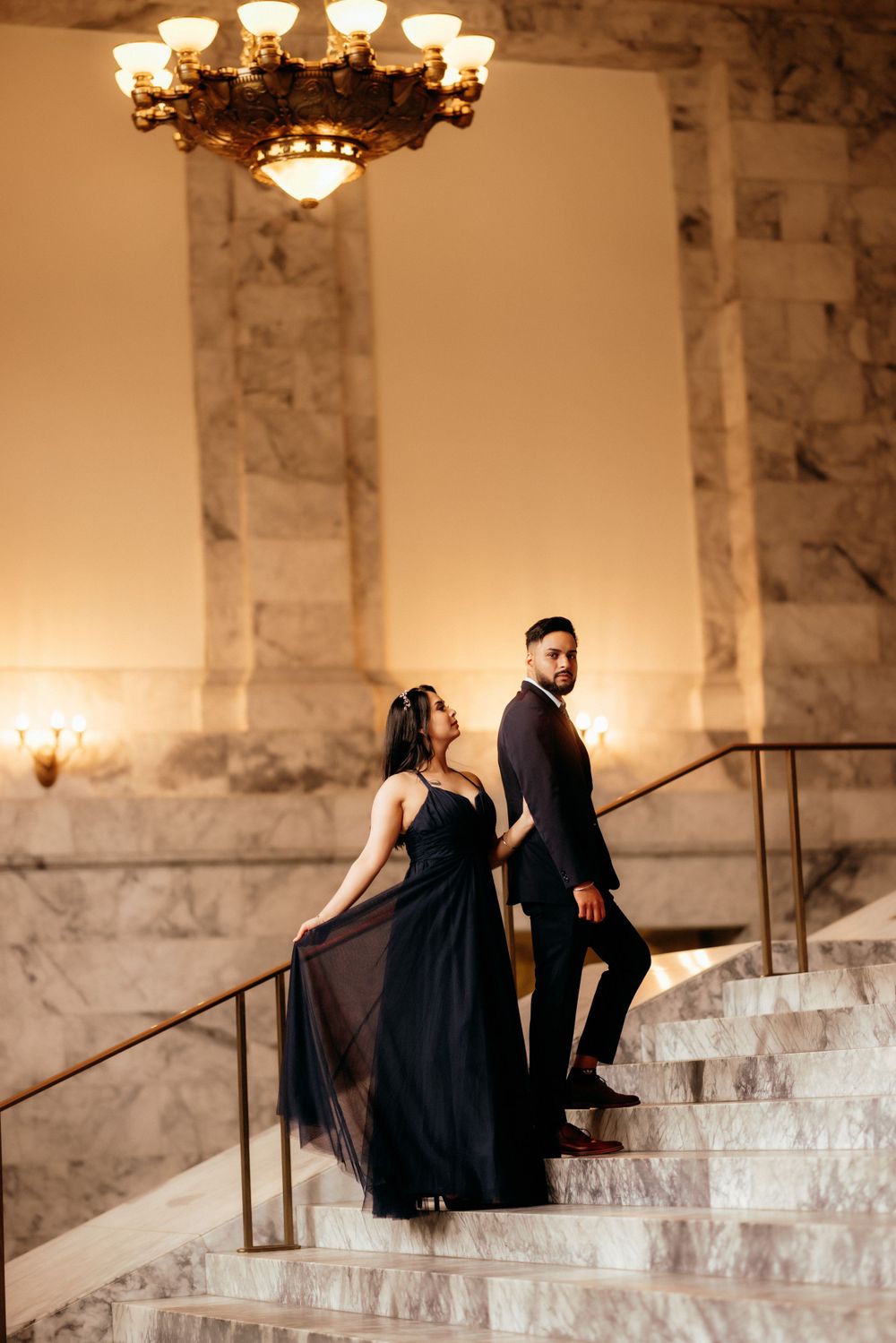 Elegant couple in formal wear posing on marble staircase under vintage chandelier.