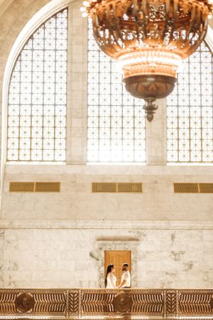 A couple stands in a grand hall with ornate chandelier and stained glass windows.