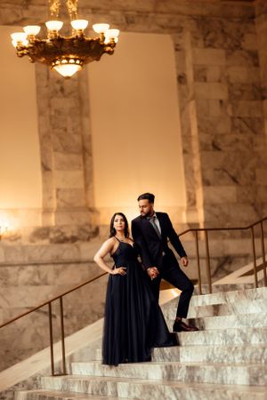 A couple in formal evening wear poses elegantly on marble steps beneath an ornate chandelier.