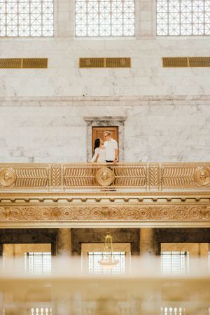 Couple poses in historic building with ornate architectural details and high windows.