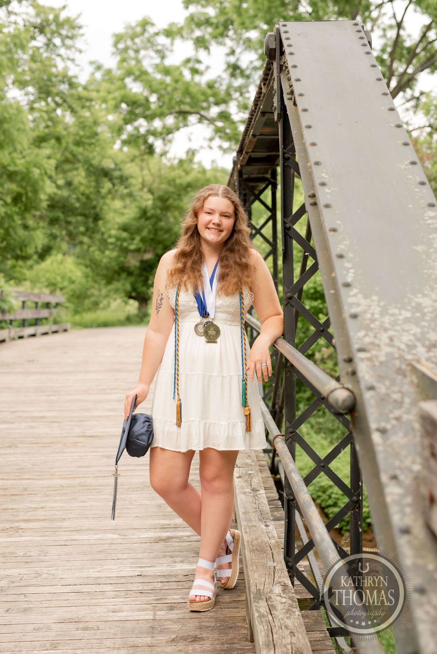 Graduate in white sundress poses with medal on wooden bridge surrounded by greenery.