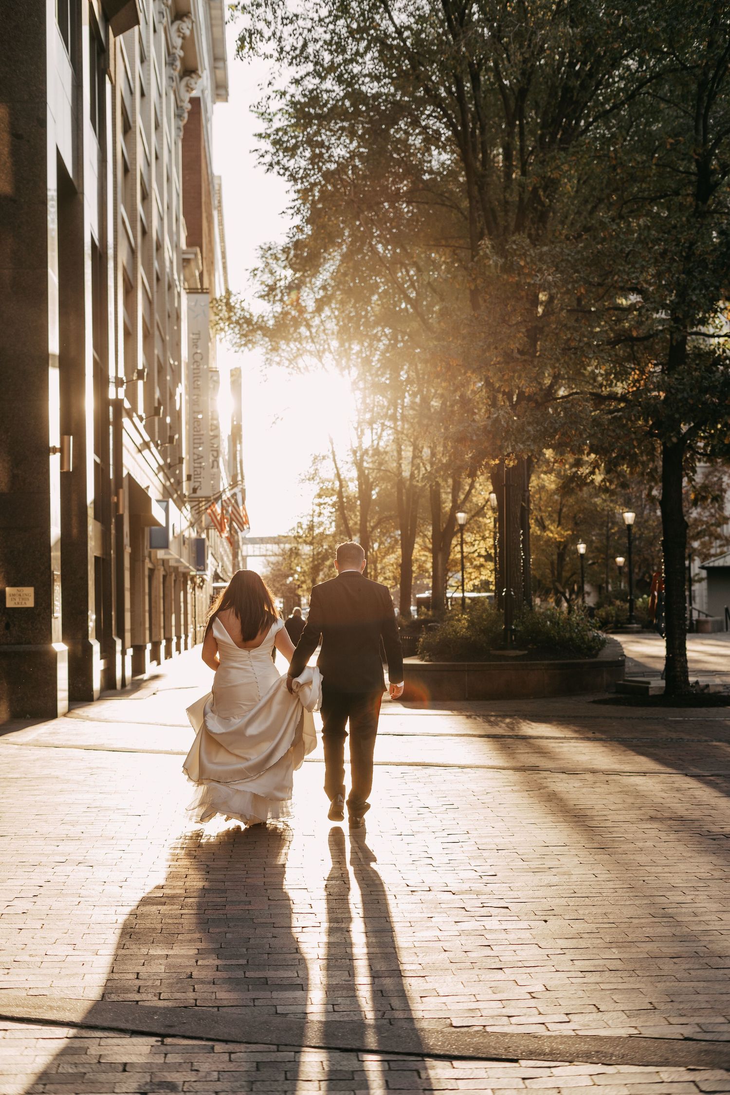 Couple walks hand in hand down sunlit urban sidewalk after wedding ceremony.