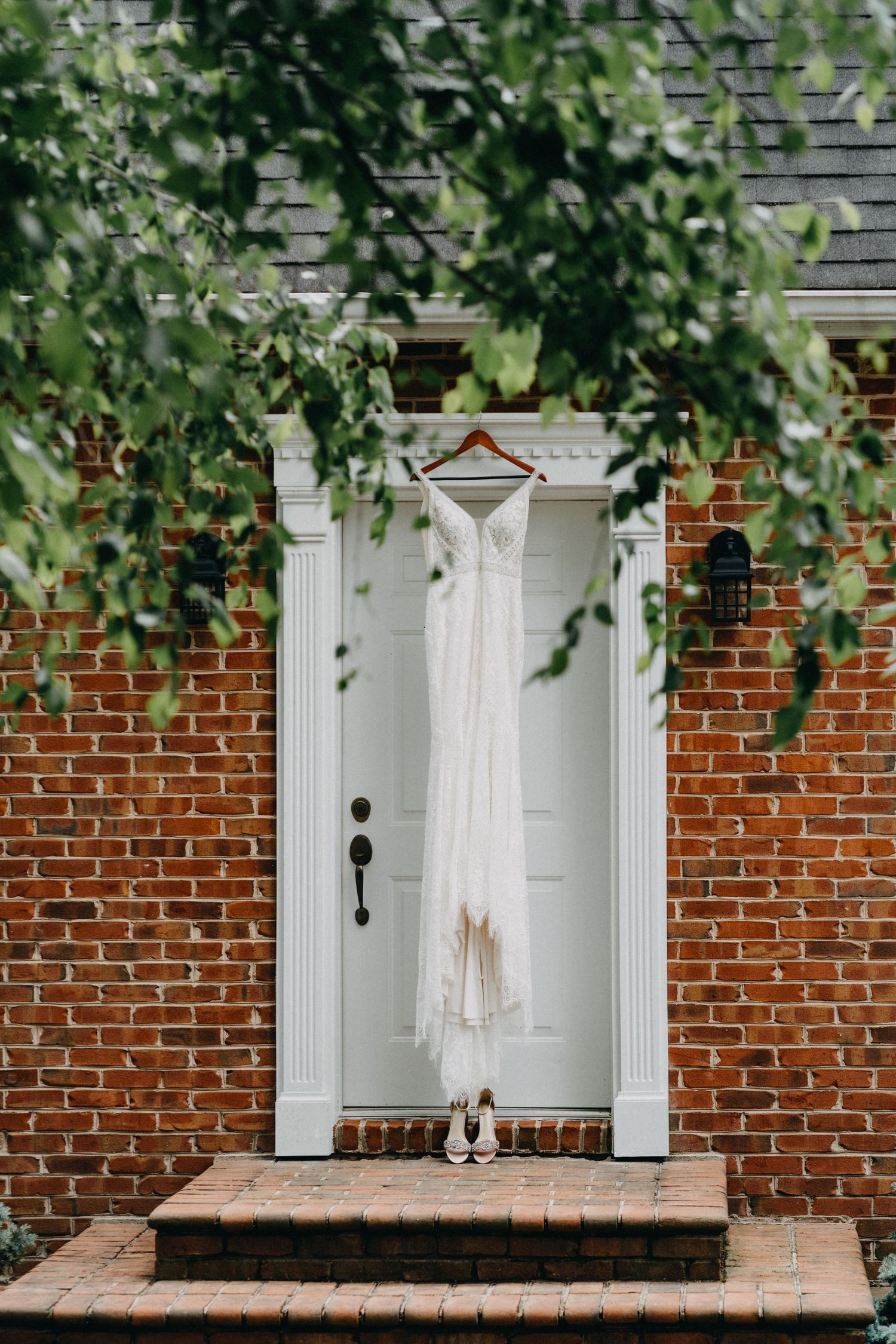 A white wedding dress hangs on a wooden hanger against a brick and white doorway with green foliage.