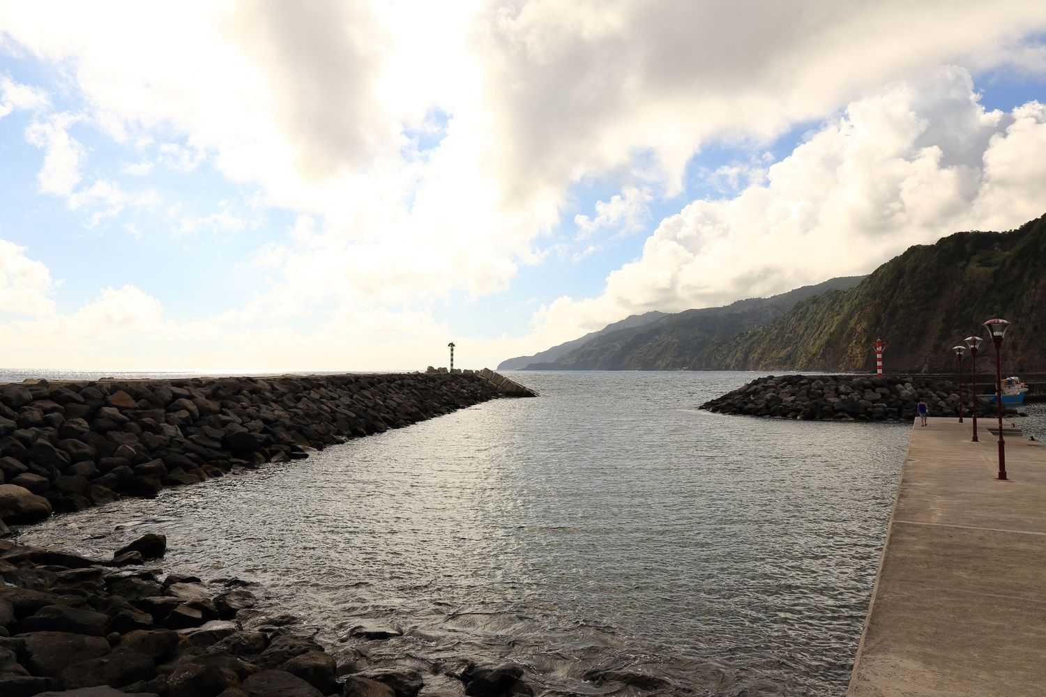 Stone breakwater extends into calm harbor waters with dramatic coastal cliffs in the background.