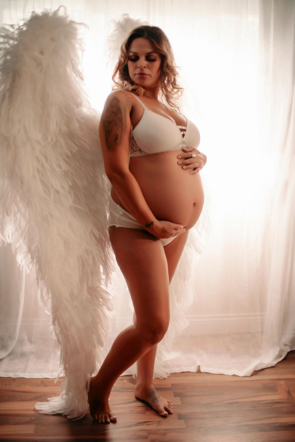 A pregnant woman in white lingerie poses with angel wings in a dreamy maternity photo shoot.