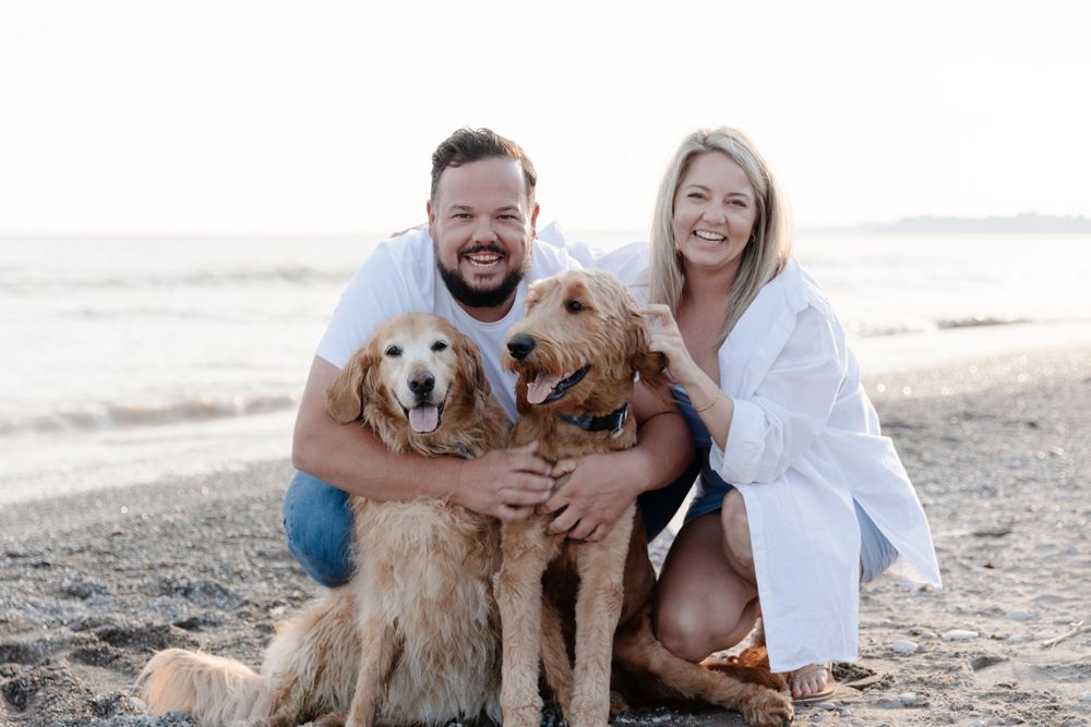 A man and woman pose with their dogs on the beach.