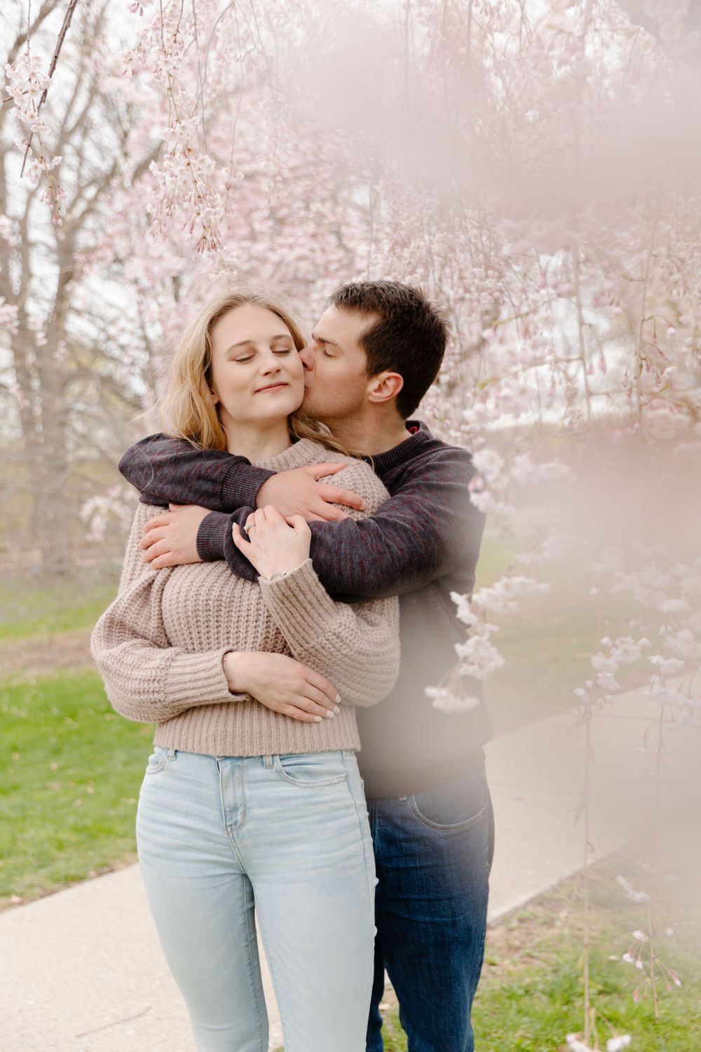 A couple kisses in front of a cherry blossom tree.