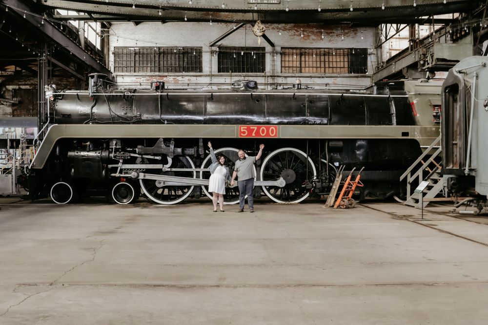 A couple standing next to an old train in a warehouse.