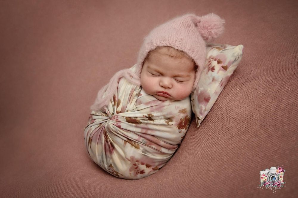 A sleeping newborn wearing a pink hat is wrapped in a floral swaddle on a dusty rose background.