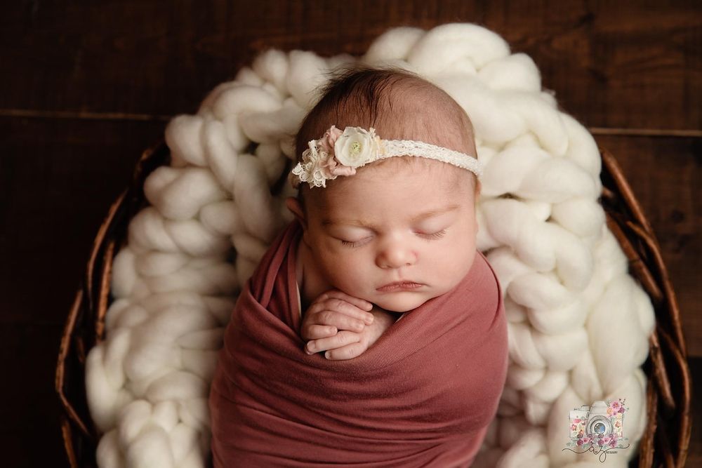 A sleeping newborn wrapped in mauve fabric rests on fluffy white wool in a wooden basket.