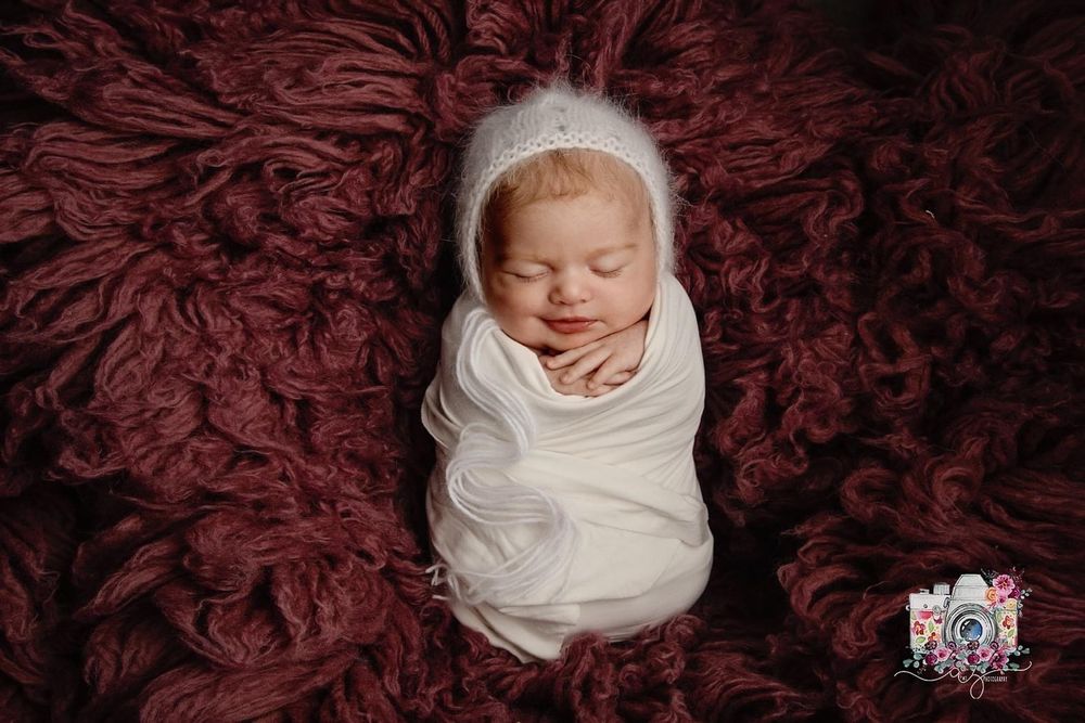 A swaddled newborn lies peacefully on luxurious burgundy textured fabric.