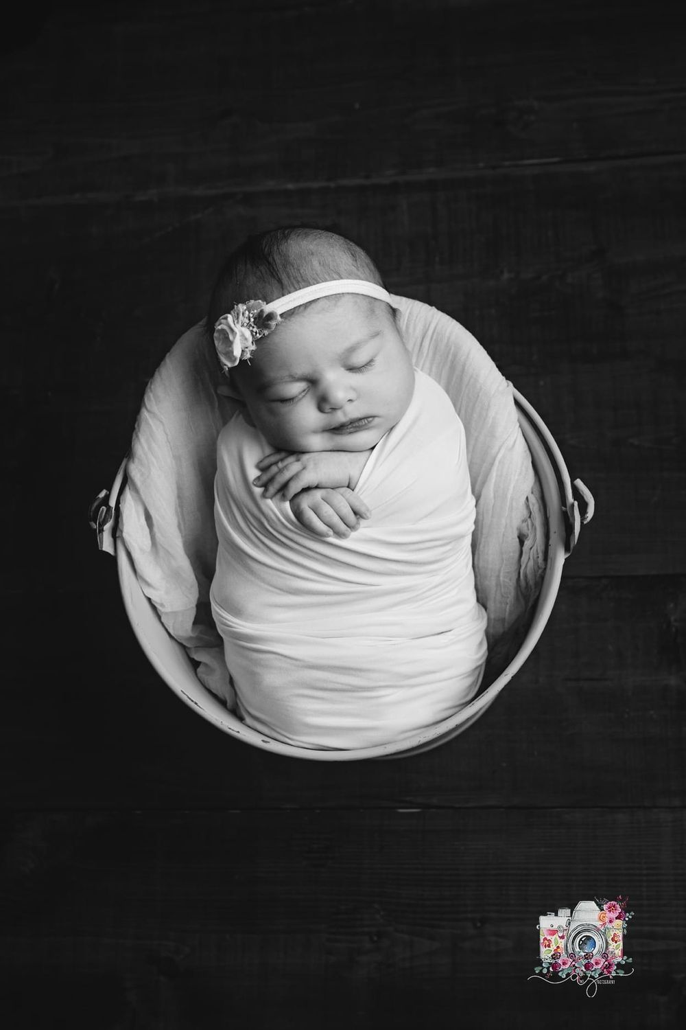 A black and white portrait of a sleeping newborn swaddled in white fabric in a round bowl.