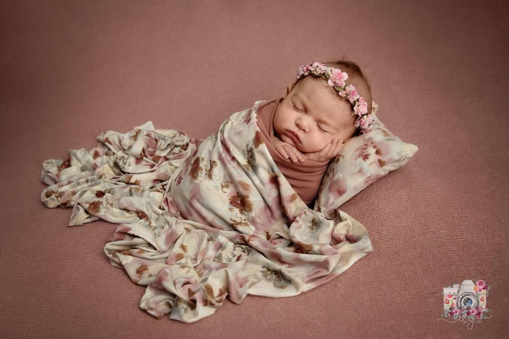 A peaceful newborn rests on a mauve backdrop wrapped in a delicate floral print blanket.