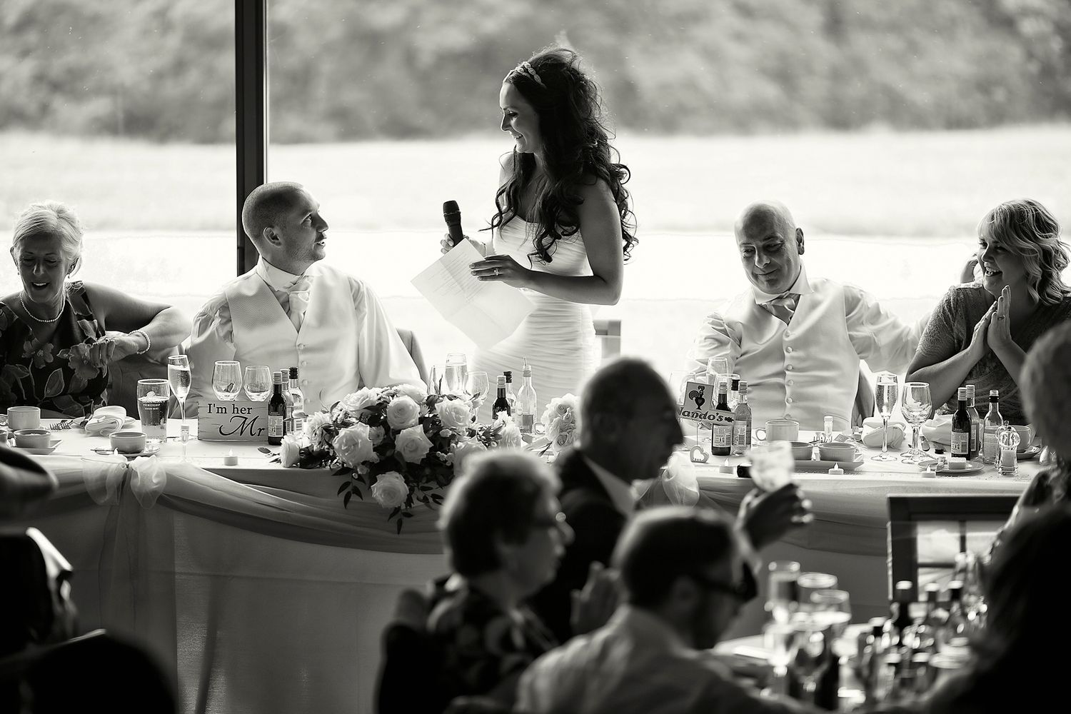 Bride gives a speech to her groom at their wedding breakfast table among family and friends