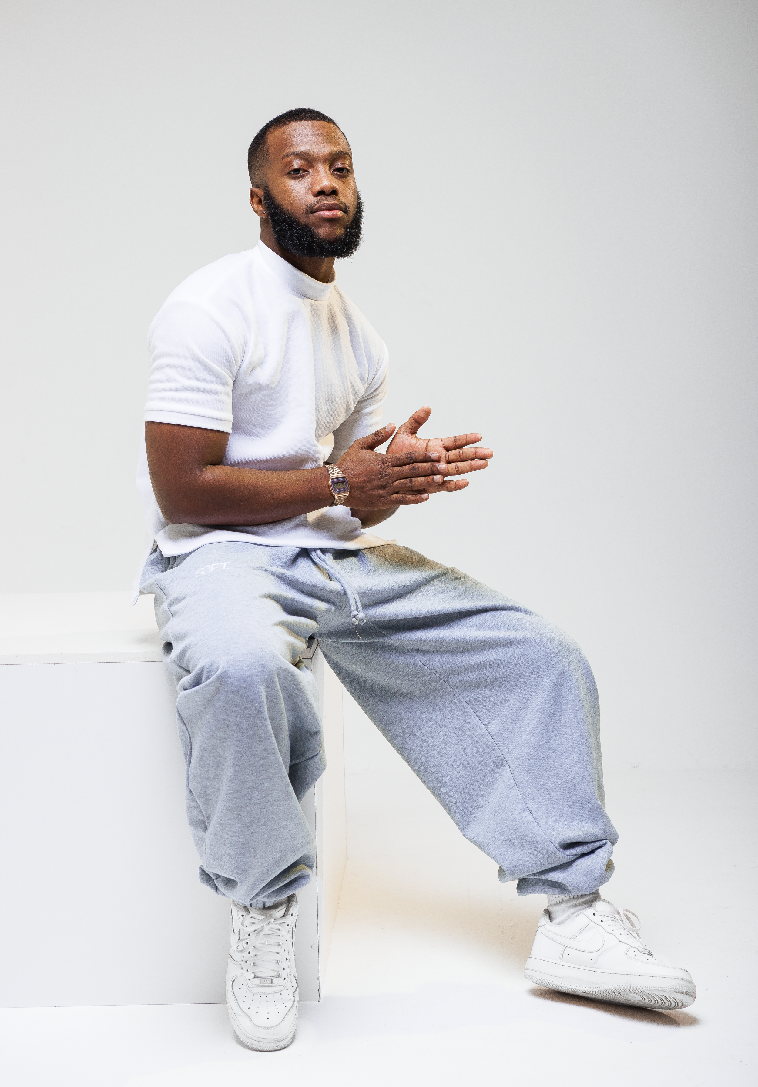 Model in white t-shirt and gray sweatpants sits casually on white platform.
