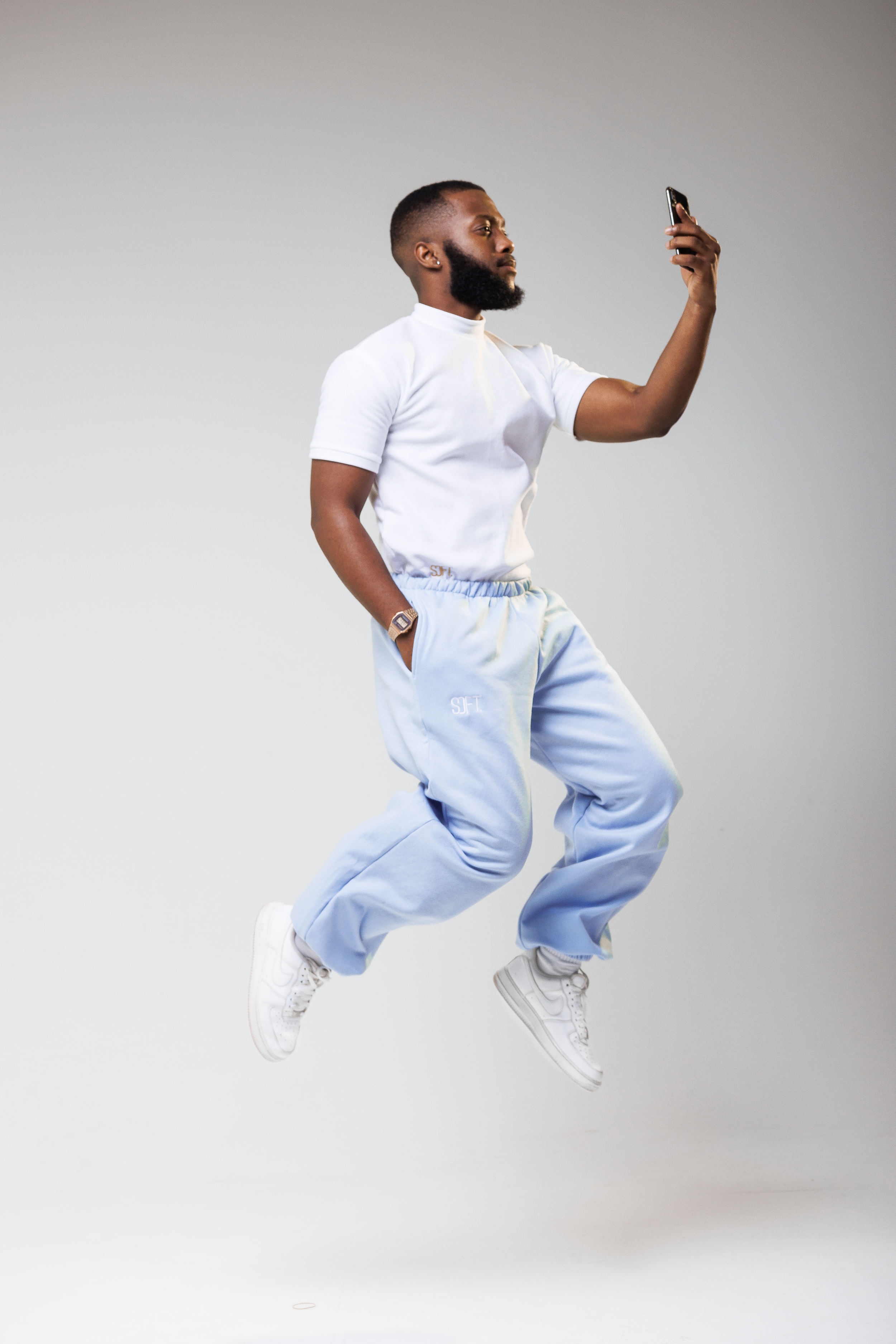 Athletic person in white t-shirt and light blue joggers jumps mid-air while looking at phone against grey background.