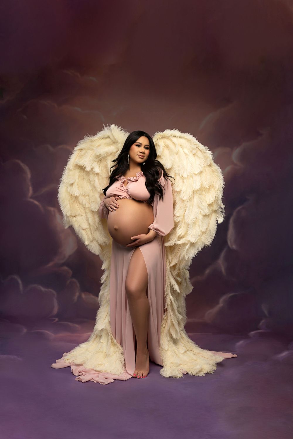 Ethereal maternity portrait featuring dramatic angel wings, flowing blush fabric, and moody purple backdrop with clouds.