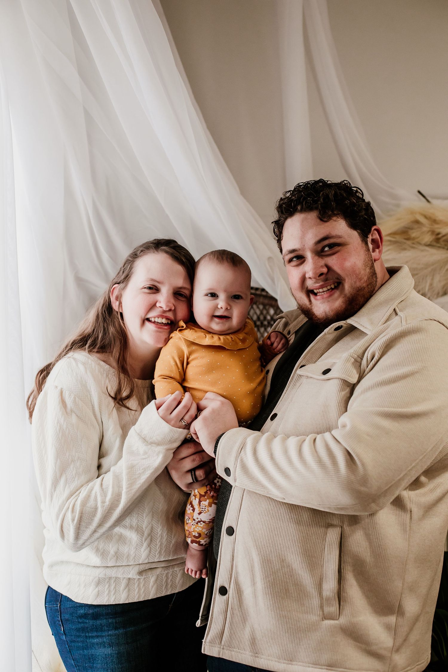 A family shares tender moments in a series of casual indoor portraits wearing cream colored clothing.
