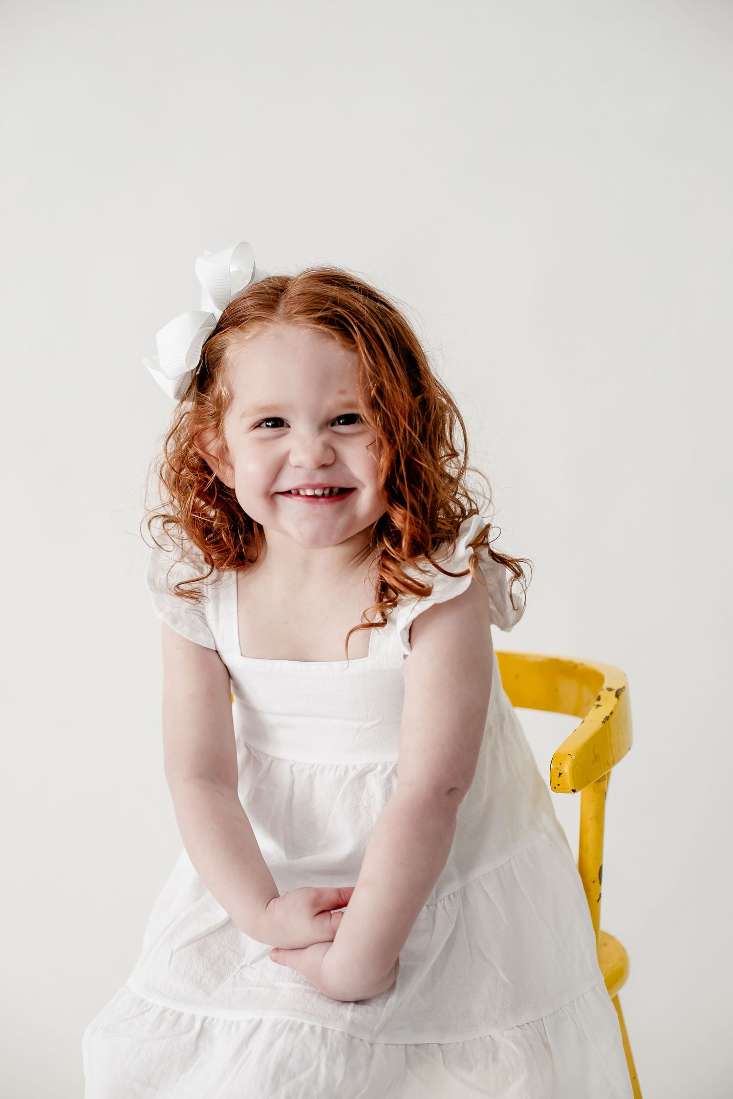 Young child in white dress sitting on yellow chair against white background.
