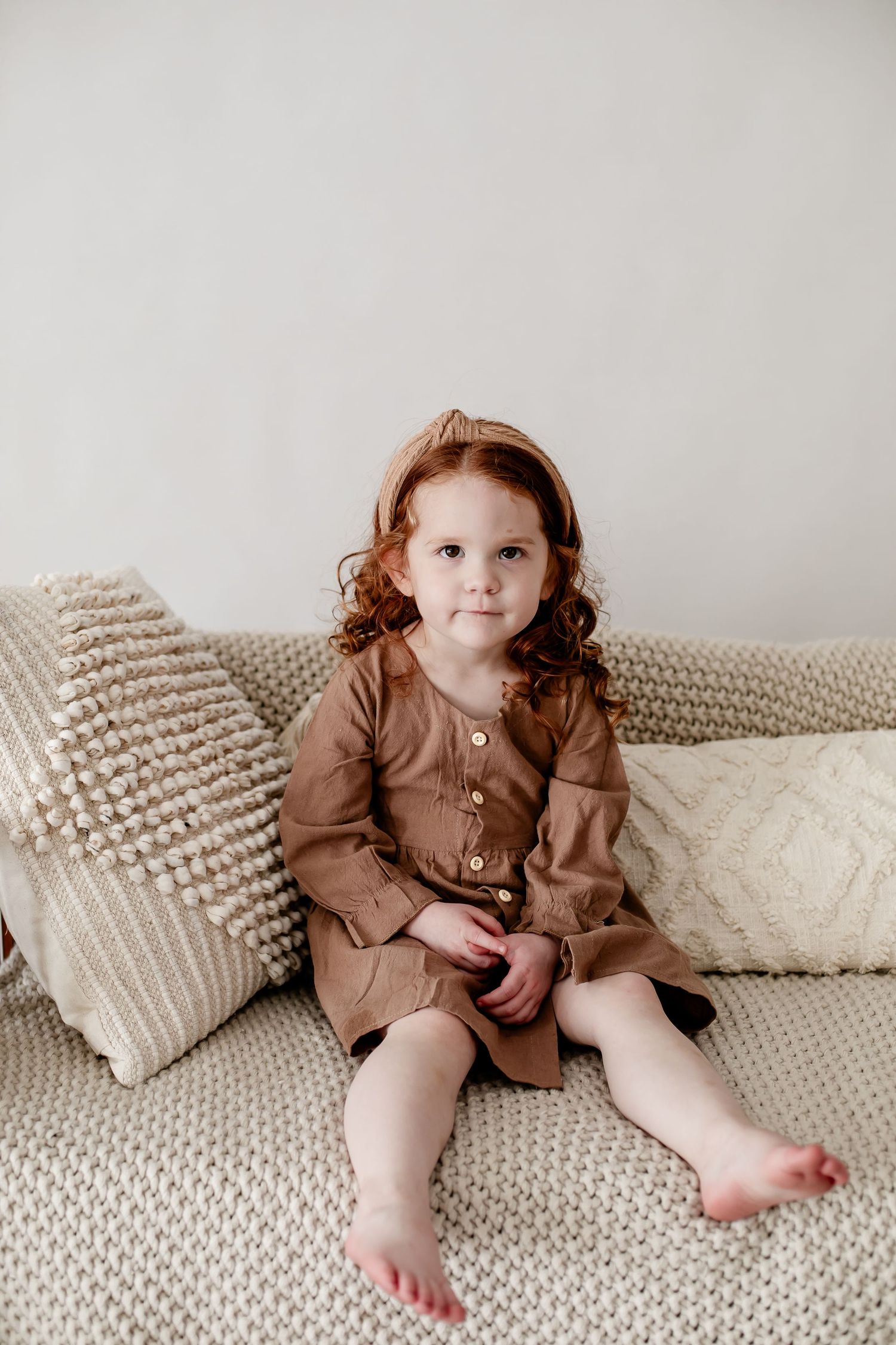 Child in brown dress sitting on textured couch with decorative pillows.