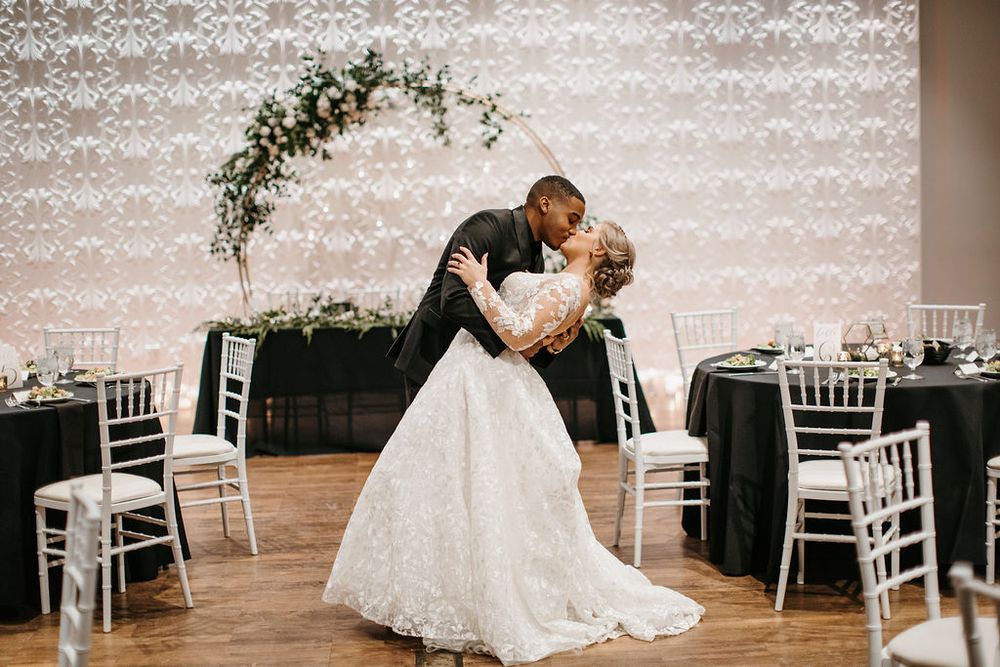 Bride and groom share romantic dip kiss in front of circular floral arch at elegant wedding reception.