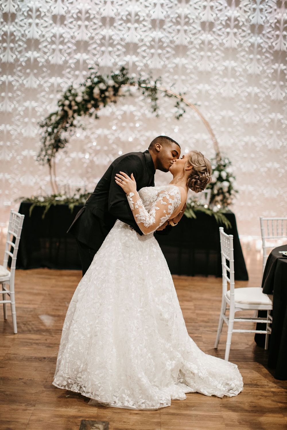 Interracial bride and groom share first dance kiss in front of floral hoop arch at wedding reception.