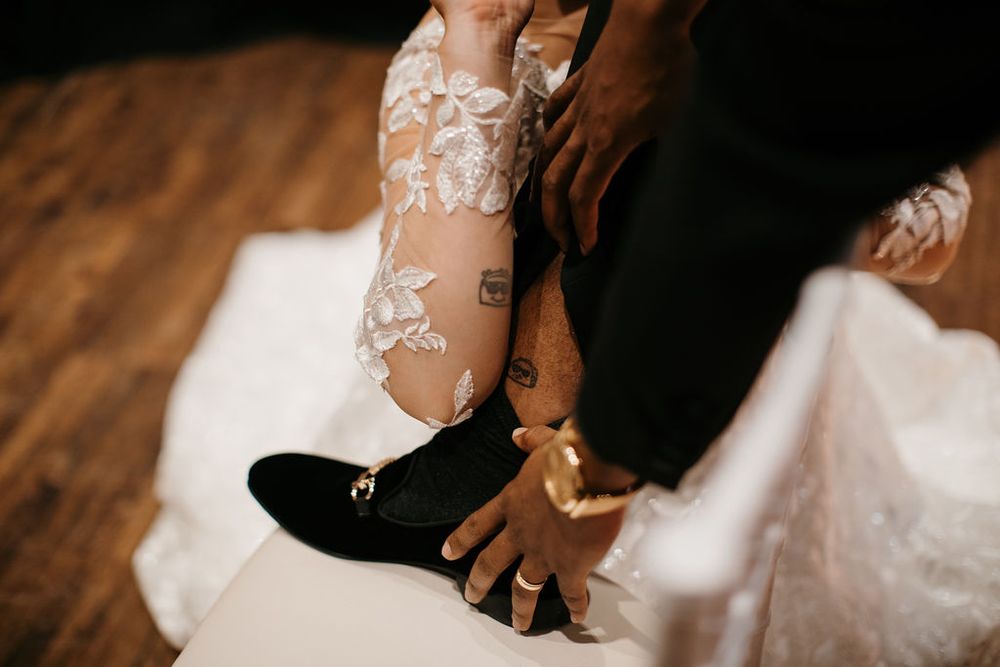 Close-up of bride's lace wedding dress and tattooed legs beside groom's black loafer shoes.