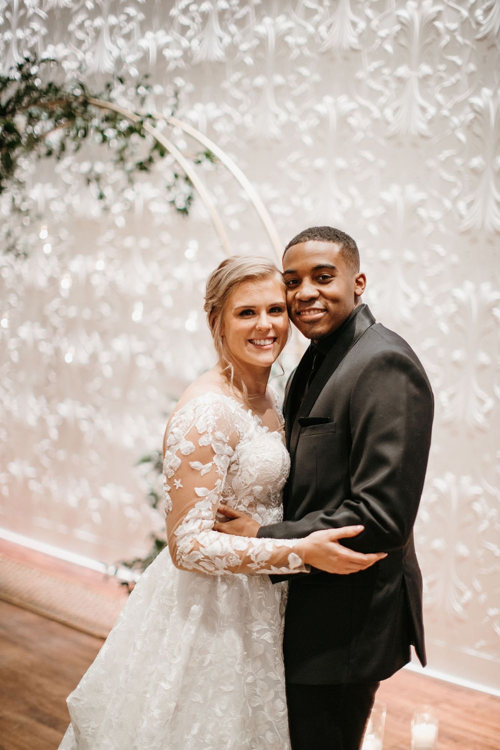 Smiling interracial bride and groom pose together in front of floral hoop arch at wedding reception.