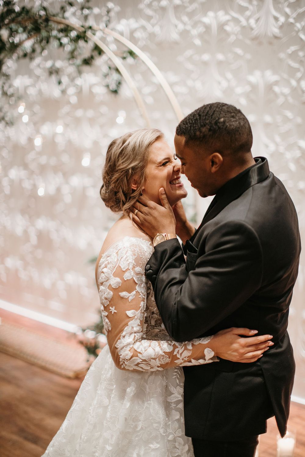 Interracial newlyweds share an intimate moment laughing together in front of decorative wedding backdrop.