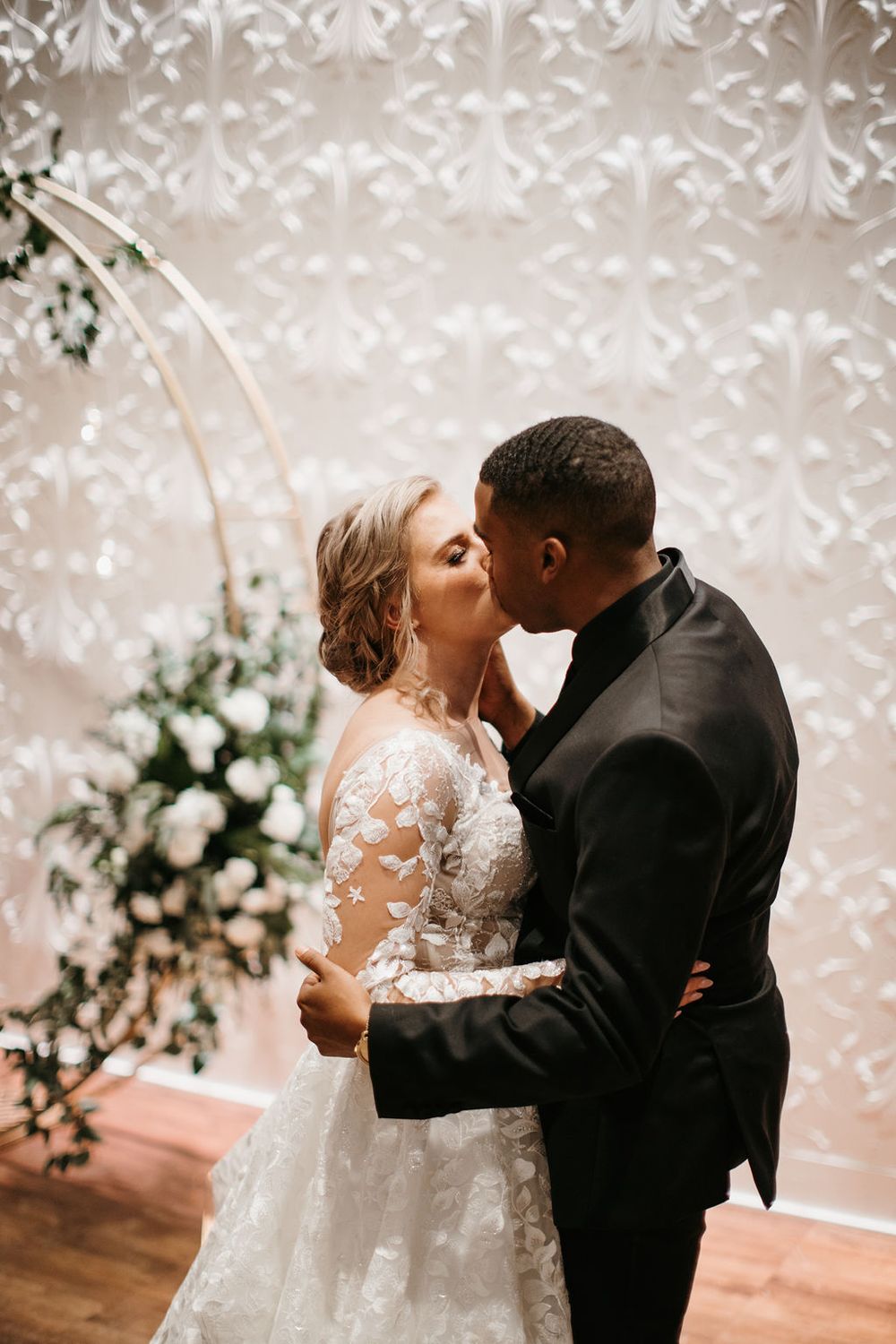 Interracial couple sharing first kiss at wedding altar with floral arch and decorative white backdrop.