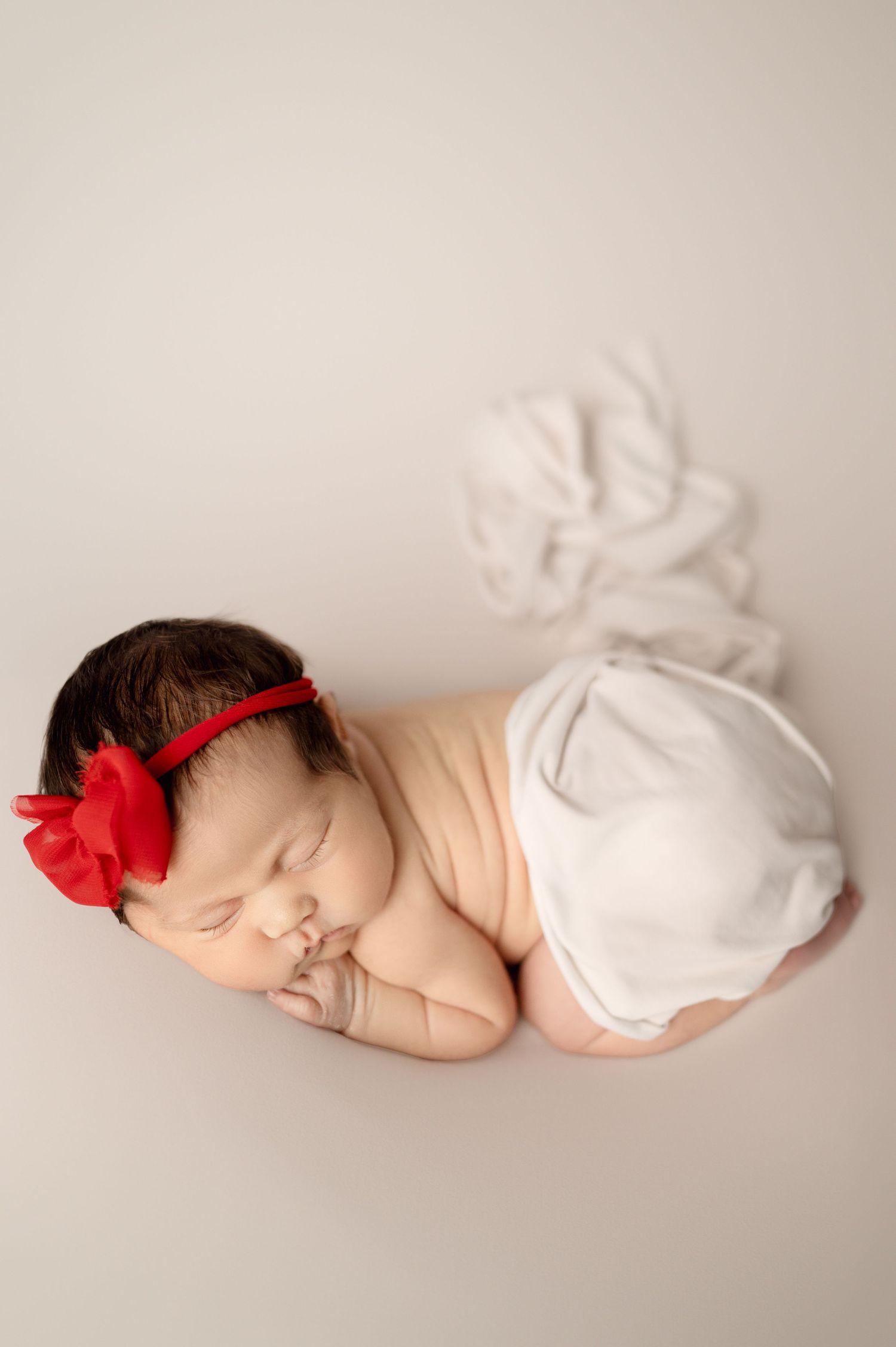 Artistic newborn portrait with red bow and white wrap on minimalist light backdrop.