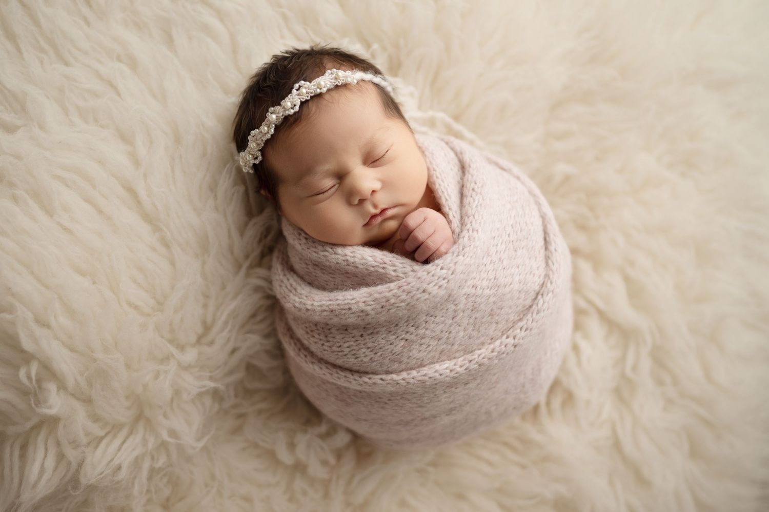 Sleeping newborn in soft neutral wraps on cream textured fur backdrop.