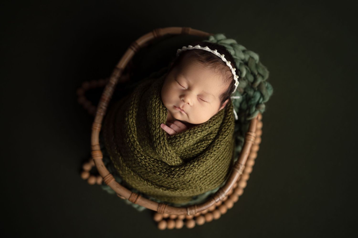 Newborn sleeping peacefully in dark green knit wrap on wooden bowl against black backdrop.