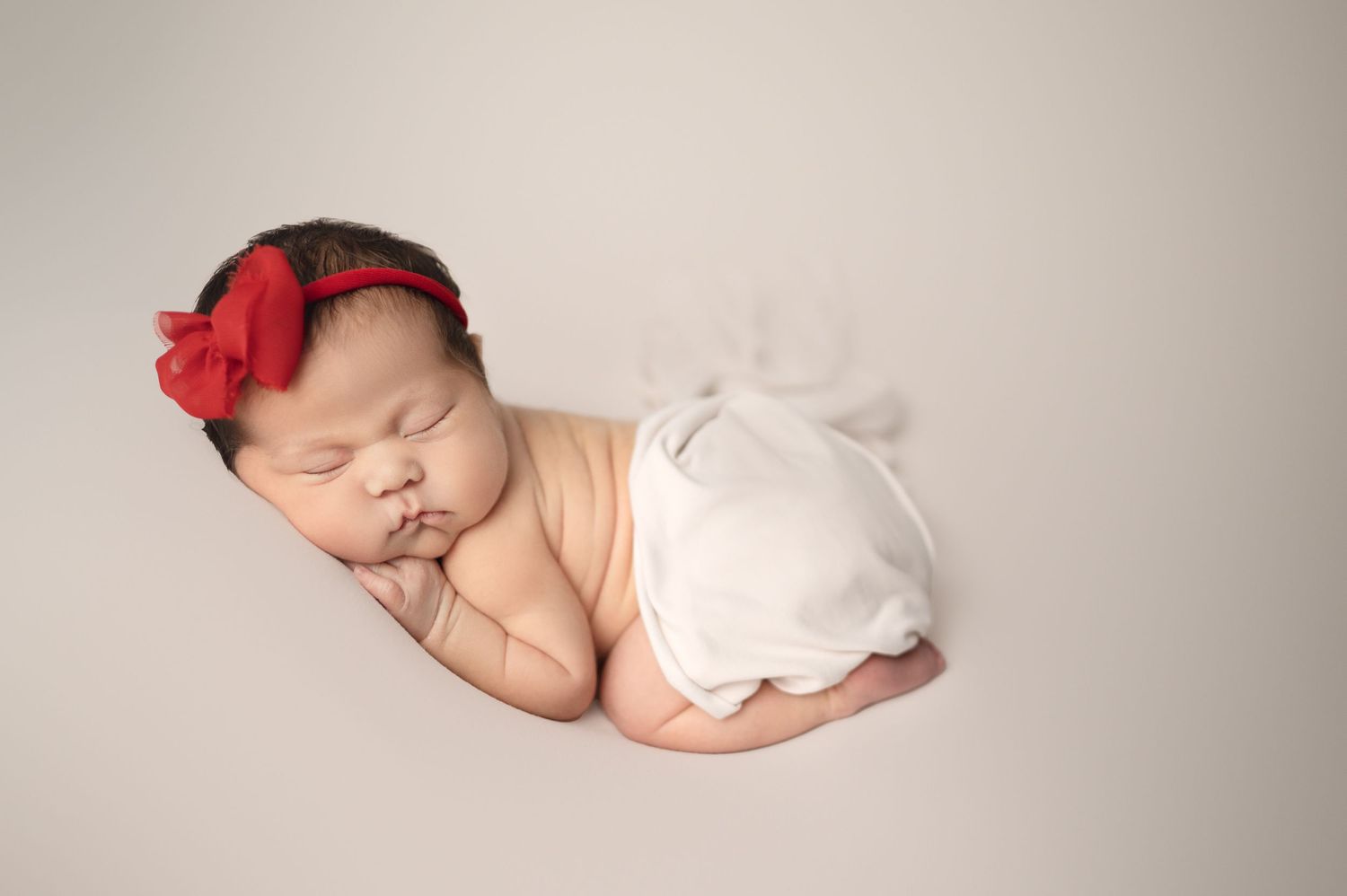Sleeping newborn with red bow accessory and white wrap on light backdrop.